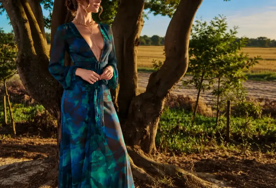 High-class escorts Agency Stockholm, Sweden: elegant escort in a blue-green print gown beside a tree at sunset