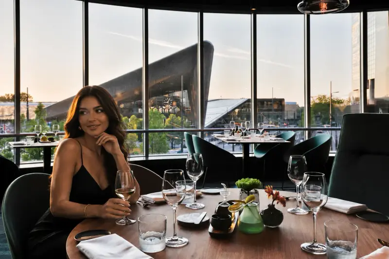 A high-class dinner date escort enjoying a glass of wine at The Millèn restaurant in Rotterdam with the city's iconic architecture in the background with Top Companions